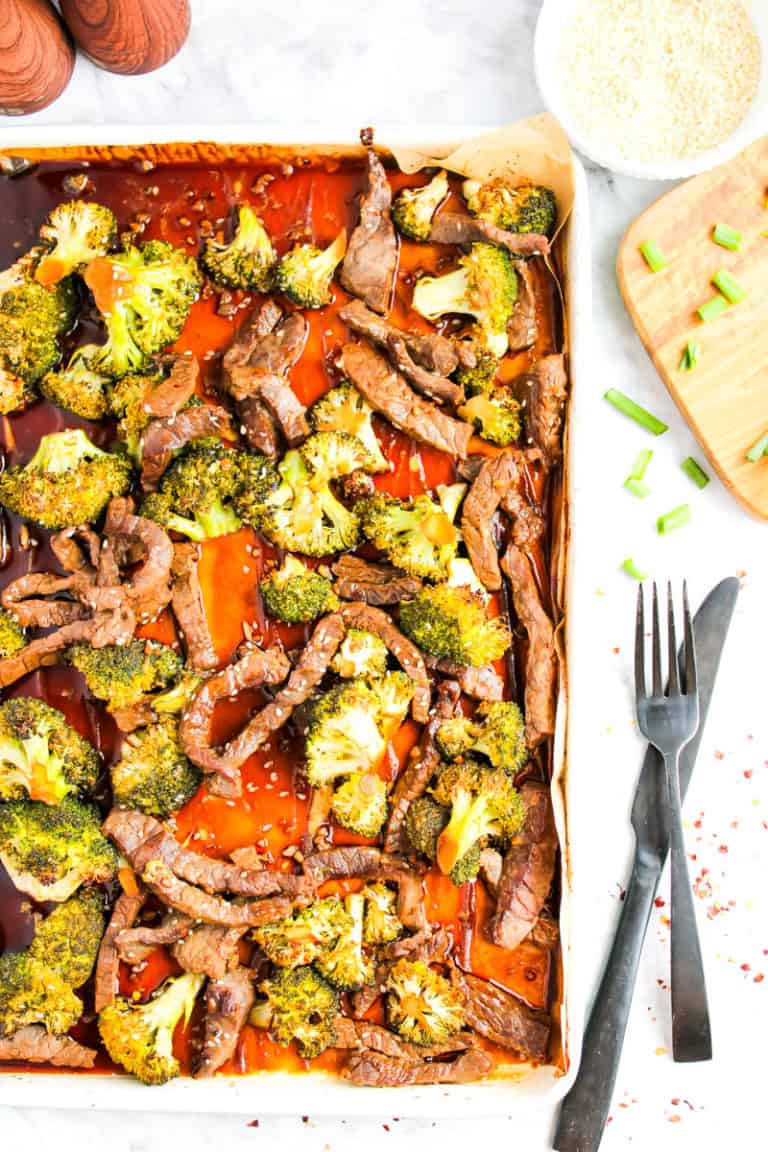 Sheet pan with beef and broccoli along with marinade sauce on white background. A silver fork and knife are next to pan. A few pieces of chopped green onion and bowl of rice in right corner of picture.