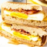 Two sandwiches stacked on top of one another. Bread, egg, bacon and top layer of bread wrapped in parchment paper with toothpick sticking in top.