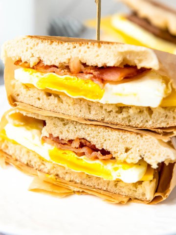 Two sandwiches stacked on top of one another. Bread, egg, bacon and top layer of bread wrapped in parchment paper with toothpick sticking in top.