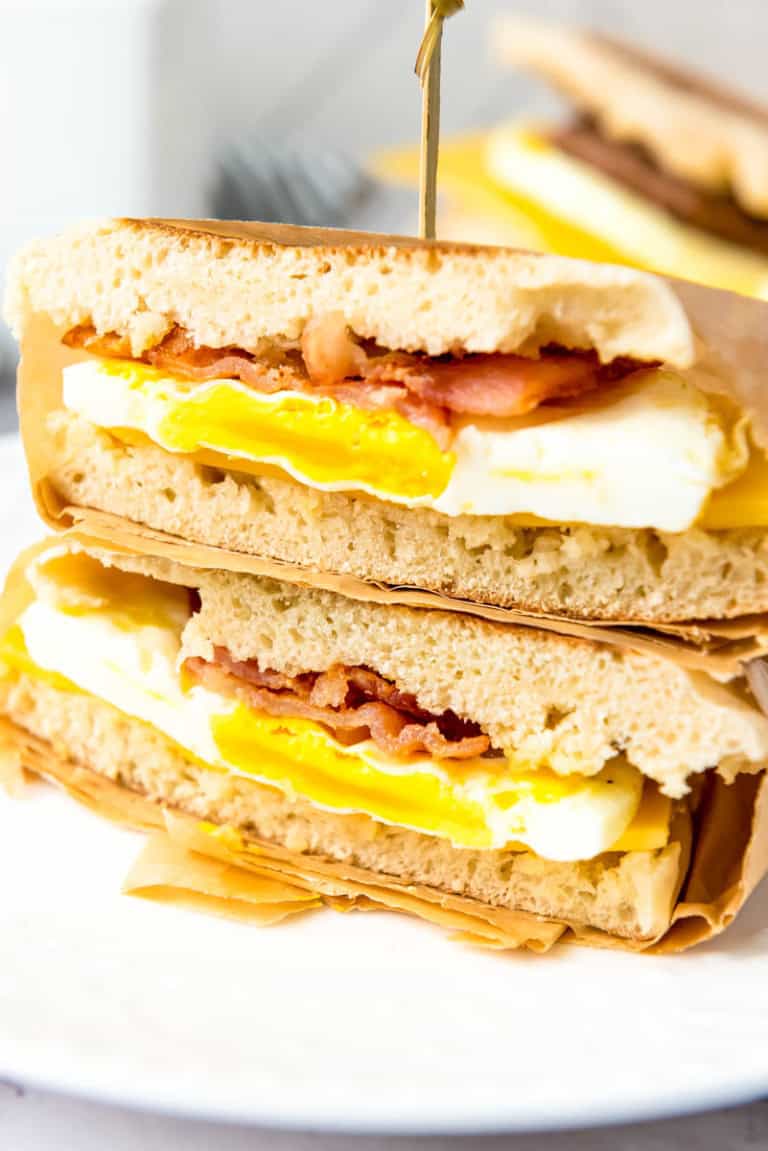 Two sandwiches stacked on top of one another. Bread, egg, bacon and top layer of bread wrapped in parchment paper with toothpick sticking in top.