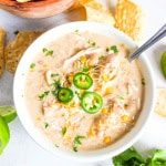 White bowl with white chicken chili with chips scattered behind along with cilantro and a lime.