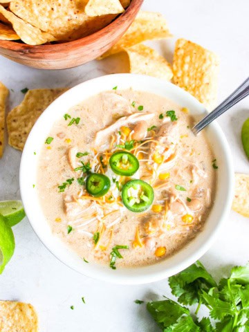 White bowl with white chicken chili with chips scattered behind along with cilantro and a lime.