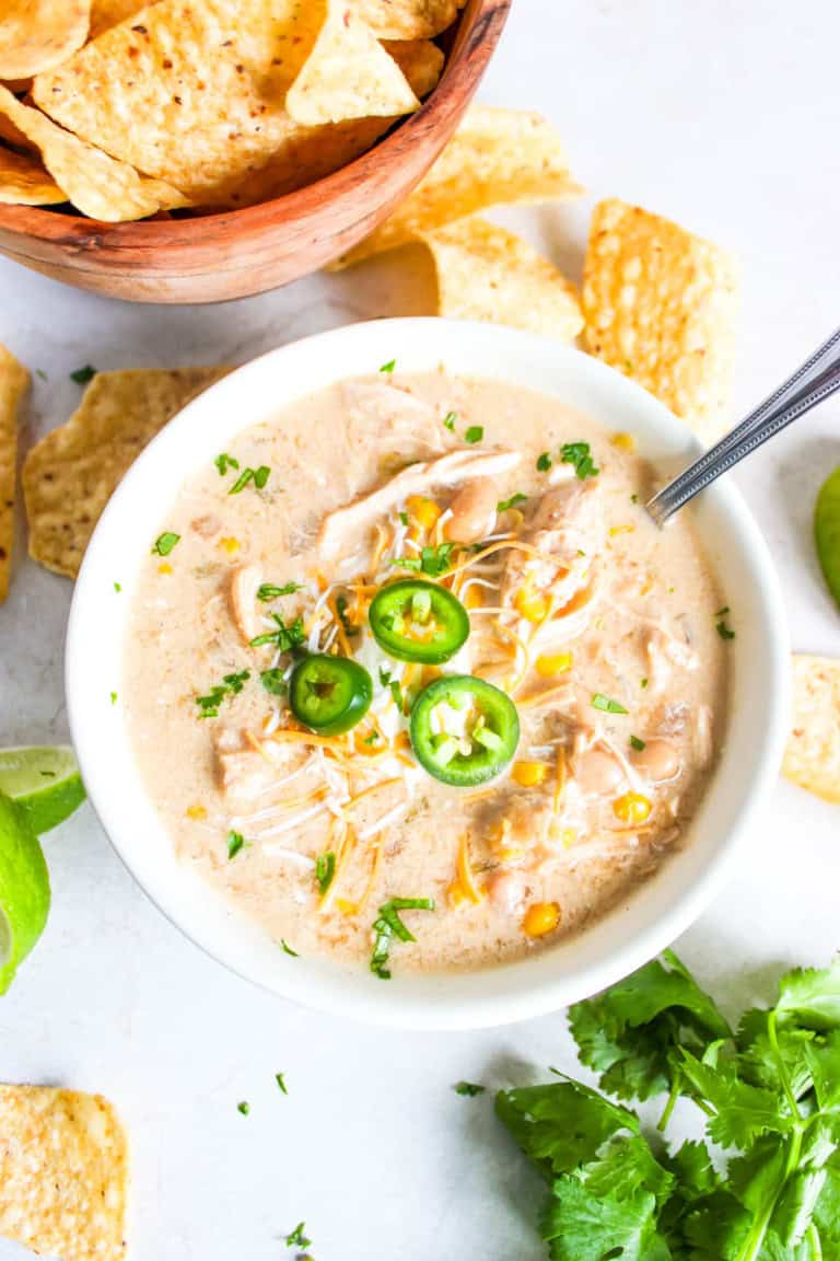 White bowl with white chicken chili with chips scattered behind along with cilantro and a lime.