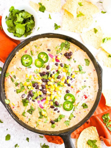 Cowboy queso dip in skillet on orange towel with chips and jalapenos in a jar around it.