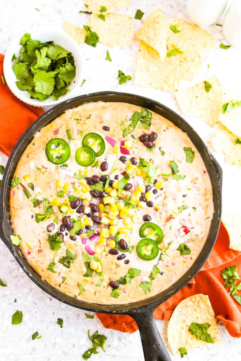 Cowboy queso dip in skillet on orange towel with chips and jalapenos in a jar around it.