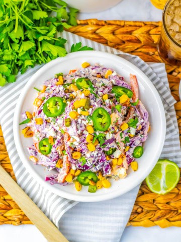 White bowl of mexican street corn coleslaw on strip towel with cilantro and a sliced lime next to the bowl.