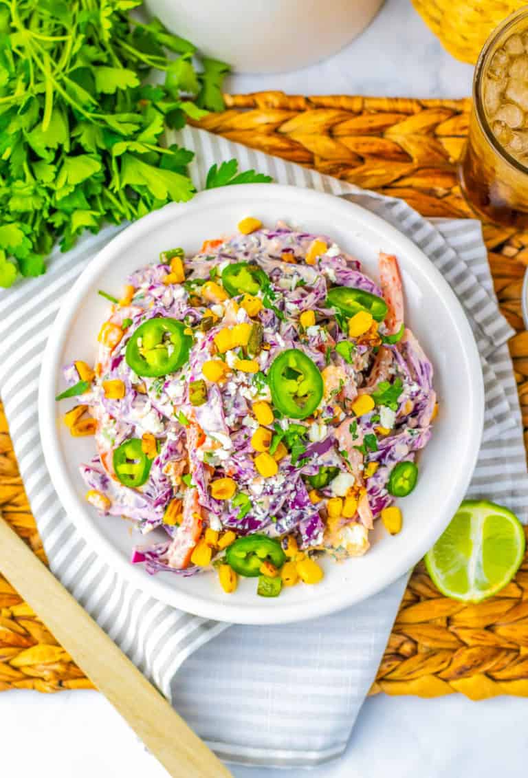 White bowl of mexican street corn coleslaw on strip towel with cilantro and a sliced lime next to the bowl.