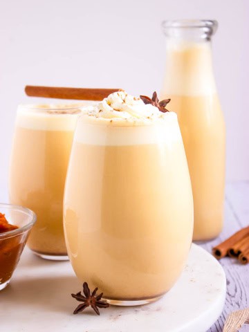 Three glasses of pumpkin spice eggnog with cinnamon sticks and pumpkin spice on the side.