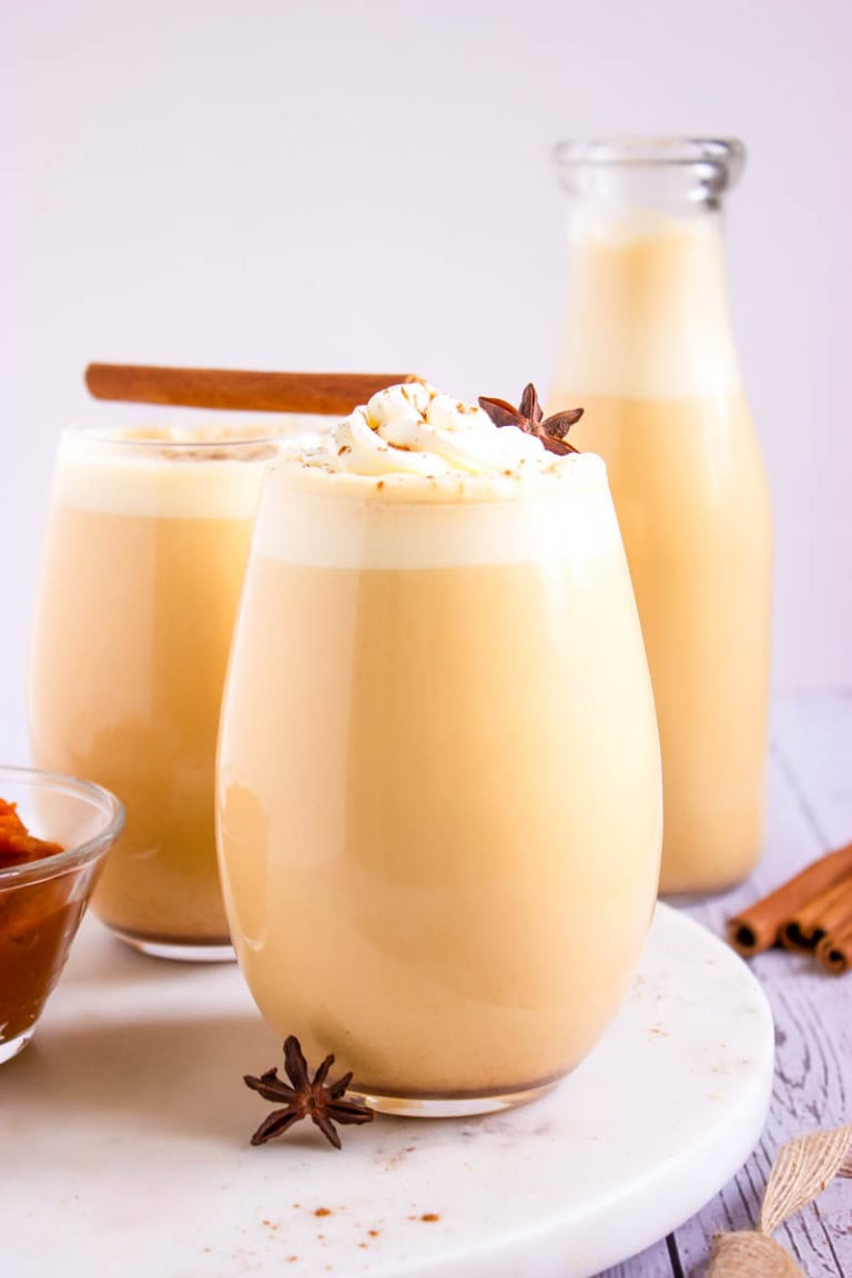 Three glasses of pumpkin spice eggnog with cinnamon sticks and pumpkin spice on the side.