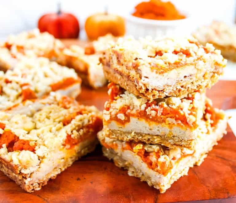 The pumpkin cheesecake crumble bars stacked on a wooden cutting board with little mini pumpkins in the background.