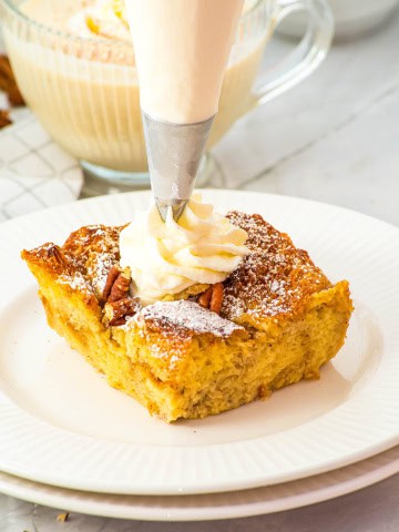 A piece of gluten free eggnog french toast casserole on a white plate with whipped cream on top of the piece.
