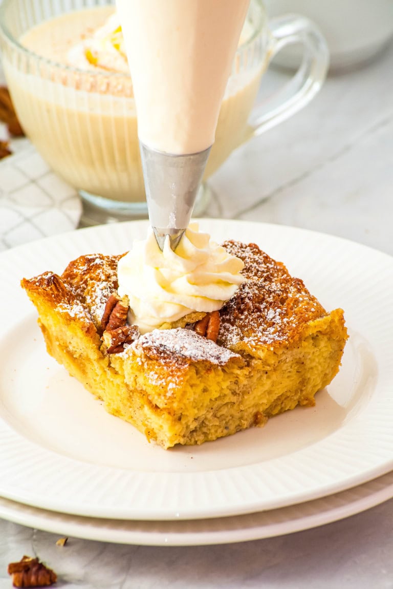 A piece of gluten free eggnog french toast casserole on a white plate with whipped cream on top of the piece.