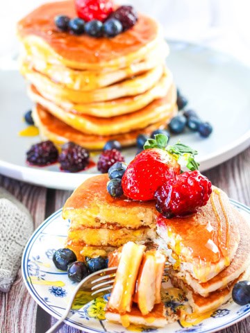 Two plates with stacks of pancakes that are topped with fresh fruit and maple syrup
