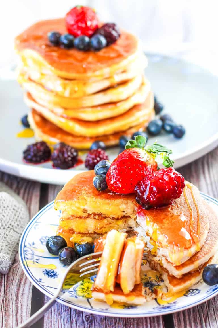 Two plates with stacks of pancakes that are topped with fresh fruit and maple syrup