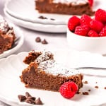 A slice of flourless chocolate torte on a white plate with another plate and slice behind it with a cup of raspberries.