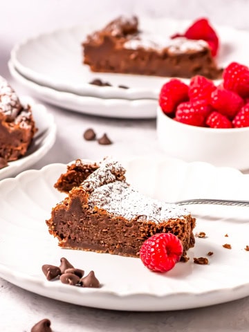 A slice of flourless chocolate torte on a white plate with another plate and slice behind it with a cup of raspberries.