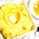 A white bowl with cheesy mashed potatoes with a plate of shredded cheese on the side with spoon.