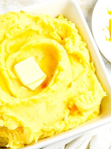 A white bowl with cheesy mashed potatoes with a plate of shredded cheese on the side with spoon.
