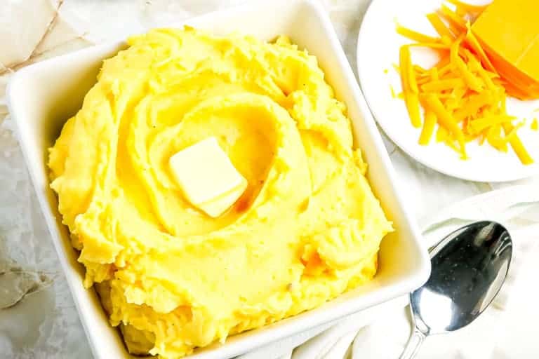 A white bowl with cheesy mashed potatoes with a plate of shredded cheese on the side with spoon.