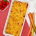 Strawberry Rhubarb crumble in white baking dish on red and white checkered towel with rhubarb and strawberries