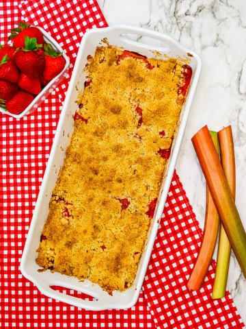 Strawberry Rhubarb crumble in white baking dish on red and white checkered towel with rhubarb and strawberries