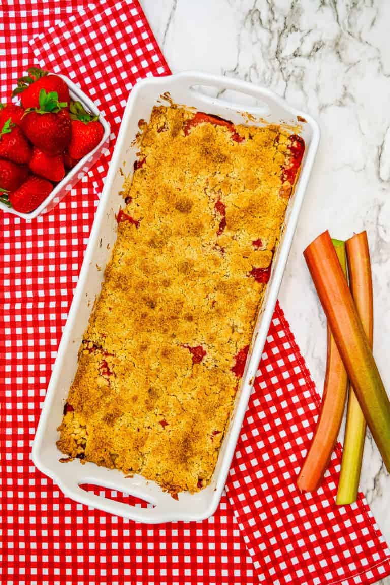 Strawberry Rhubarb crumble in white baking dish on red and white checkered towel with rhubarb and strawberries