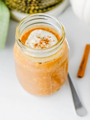 Pumpkin overnight oats in jar topped with whipped cream with cinnamon sticks on the side.