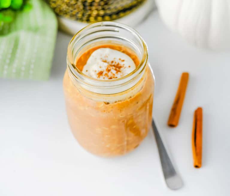 Pumpkin overnight oats in jar topped with whipped cream with cinnamon sticks on the side.