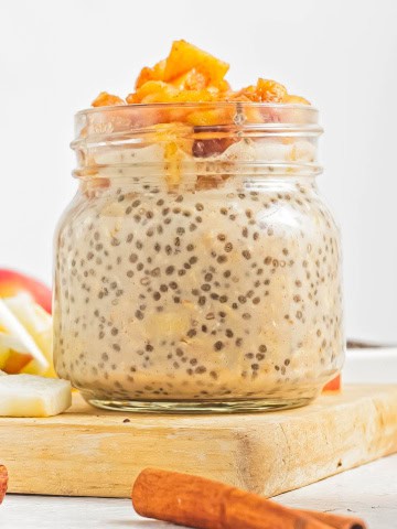 A glass jar with apple pie overnight oats sitting ontop of a piece of wood with apple pieces behind.