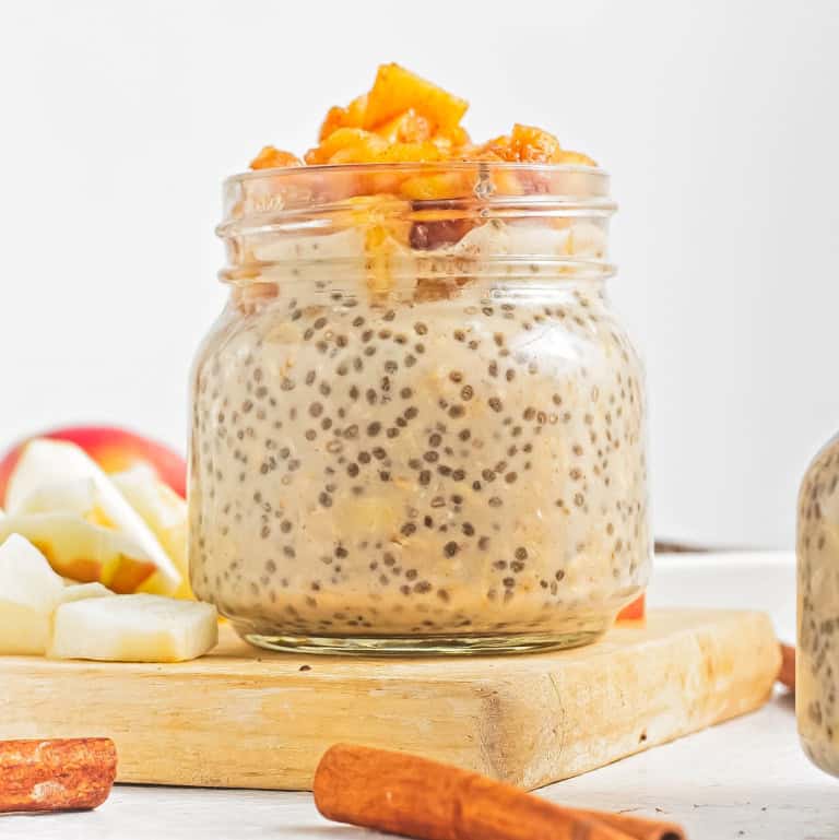 A glass jar with apple pie overnight oats sitting ontop of a piece of wood with apple pieces behind.